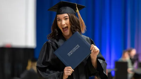 Smiling female student with dark hair wearing black graduation cap and gown, holding a diploma